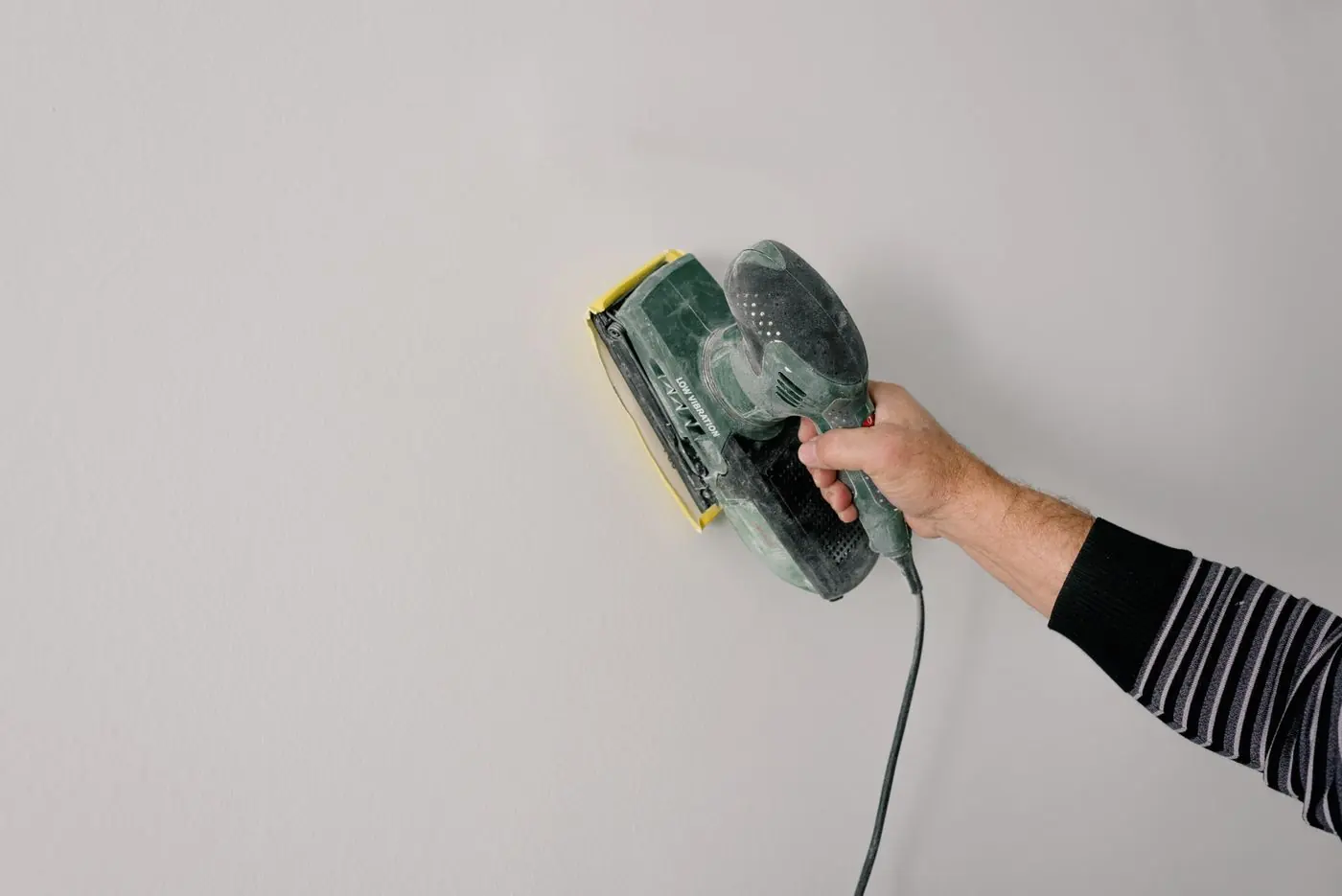 Hand schuurt een witte muur met een schuurmachine